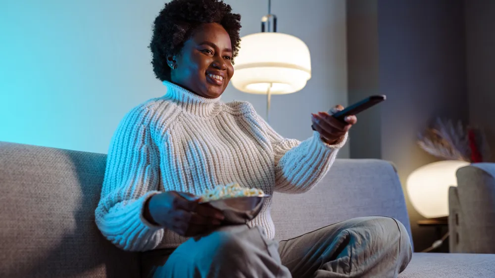 ALT Text: Cheerful smiling woman is watching TV and holding the remote control. Cozy night at home